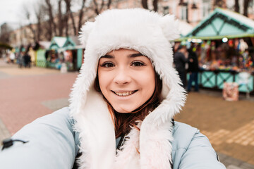 Young pretty smiling woman celebrating dressed warm jacket and woven woolen hat, taking sealfie with a phone. Winter market holidays fair, travel spirit resort. New Year Christmas decorations

