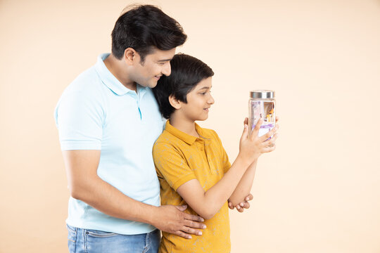 Portrait Of Happy Young Indian Father And Son Holding Glass Jar Full Of Rupee Notes Isolated On Beige Studio Background. Saving Money And Investing Concept.