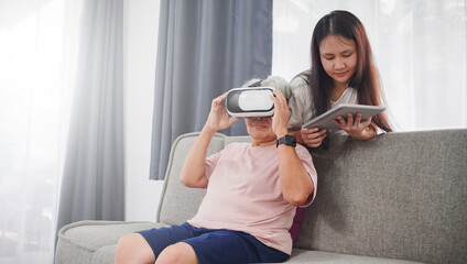Virtual reality and metaverse at home concept, senior asian woman wearing vr glasses, with her daughter controlling using tablet, in living room at home.