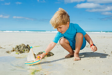 Handsome little blond boy put toy boat in paddle on sea beach