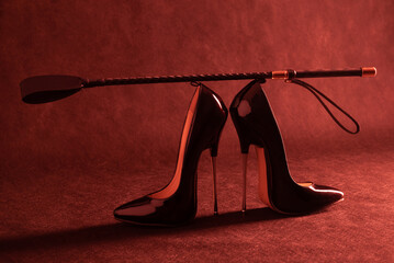 Black shiny high heel shoes and a whip on a dark background - obrazy, fototapety, plakaty