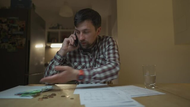 Man argues over phone after getting utility bill in evening at home sitting at table in kitchen. Male swearing on phone looking at bank receipt calculating taxes, examining an invoice document. 