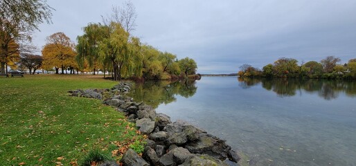 Obraz premium Calm water pond at Kingsbridge Park with autumn trees in the background on a cloudy day
