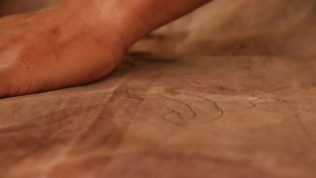 This close-up video shows a deformed adult pira's hand making a floral pattern, using a wooden pencil, in the process of making Indonesian hand-drawn batik.