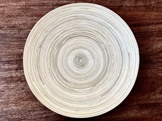 Close-up of a white wooden plate on the surface of a dark wooden tabletop. A wooden plate with a texture of concentric circles on a dark brown wooden table.