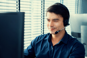Young competent asian male call center agent working at his computer while simultaneously speaking...