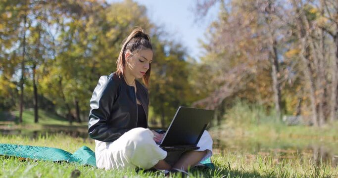 Beautiful girl working on a laptop computer while sitting on a meadow. Fashionable young lady in the park. Right camera movement.