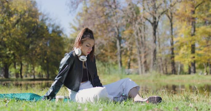 Beautiful girl sitting on a meadow in the park. Fashionable young lady with white headphones.