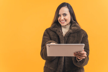 happy young Latin-American woman in a winter coat holding a tablet, isolated on orange background medium shot technology and businesswoman co cept. High quality photo