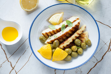 Plate with slices of grilled haloumi cheese, green olives and lemon wedges, high angle view on a light-beige stone background