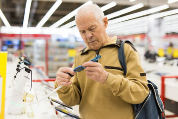 elderly grayhaired man pensioner examining counter with electronic gadgets and smart watches in showroom of digital goods store