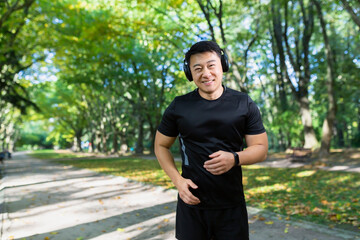 Portrait of a young Asian athlete running in the park, forest, listening to music, podcasts in headphones. He smiles, looks at the camera.