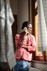 Smiling millennial African guy talking on phone, standing by window at home, happy young black man holding smartphone answering call, speaking by mobile with girlfriend. Distant relationships concept