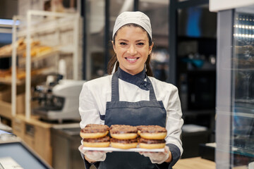 A happy baker is holding tray with doughnuts and offering them to a customers while standing at...