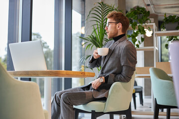 Attractive confident man holding cup of coffee looking away sitting in cafe. Coffee break concept