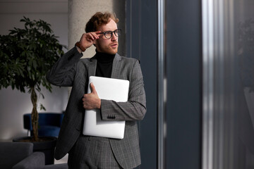 Young confident man hipster wearing stylish suit, correct eyeglasses holding laptop looking at window