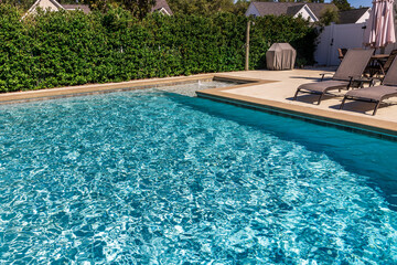 A rectangular new swimming pool with tan concrete edges in the fenced backyard of a new...