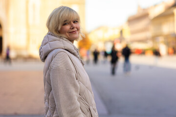 Fototapeta premium Portrait of Caucasian adult smiling blonde woman in jacket posing in city centre. Concept of carefree and psychology