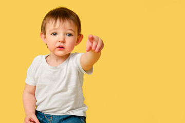 Portrait of upset toddler baby on studio yellow background. Offended child points forward with index finger in white t-shirt and blue jeans, copy space. Kid aged one year and four months