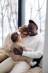 Happy interracial family sits on windowsill and plays with their baby daughter against background of window. Concept of interracial family and unity between different human races. baby daughter.