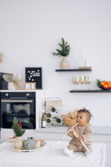 Fototapeta na wymiar Cute mixed race baby girl sits on table in kitchen and plays by wooden spoons near Christmas decoration.