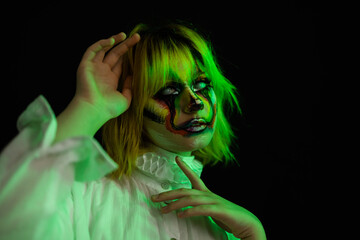 Portrait of woman with yellow hair and evil clown face art posing on black background, green side lighting