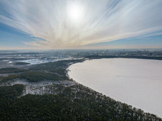 Snow-covered forest on lake shore with ice at sunset and the city on horizon, auerial view