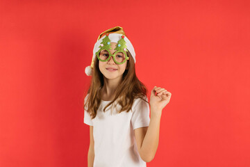 Close-up portrait of a teenage girl in a Santa cap and funny Christmas glasses on a red background, copy space