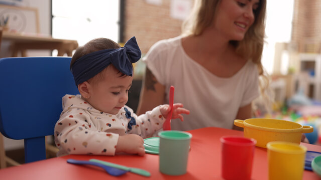 Woman and toddler learning to eat with plastic dish toy sitting on table at kindergarten