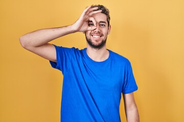 Hispanic man with beard standing over yellow background doing ok gesture with hand smiling, eye looking through fingers with happy face.