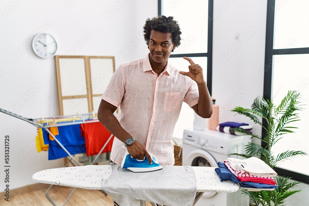 Wall mural african man with curly hair ironing clothes at home smiling and confident gesturing with hand doing 