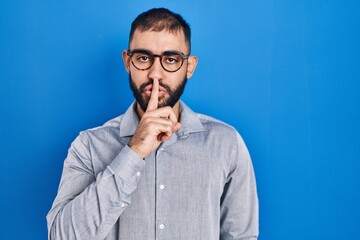Middle east man with beard standing over blue background asking to be quiet with finger on lips. silence and secret concept.