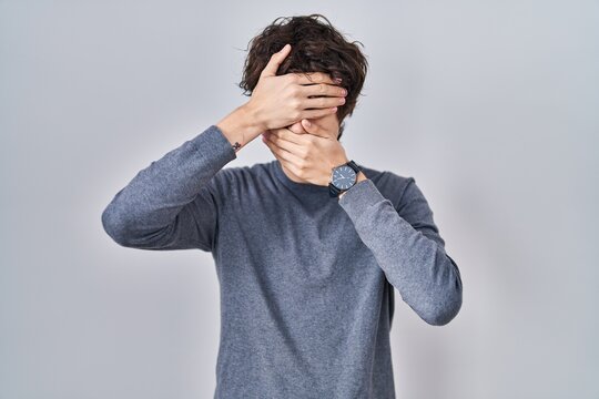 Young man standing over isolated background covering eyes and mouth with hands, surprised and shocked. hiding emotion