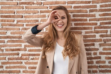Beautiful blonde woman standing over bricks wall doing ok gesture with hand smiling, eye looking through fingers with happy face.