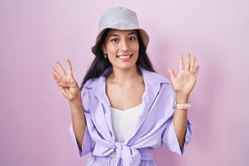 Obraz na płótnie Canvas Young hispanic woman standing over pink background wearing hat showing and pointing up with fingers number nine while smiling confident and happy.