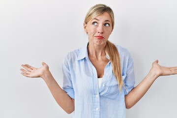 Beautiful blonde woman standing over white background clueless and confused expression with arms...
