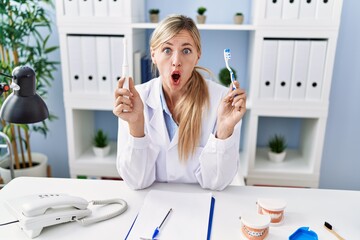 Beautiful dentist woman holding ordinary toothbrush and electric toothbrush in shock face, looking...