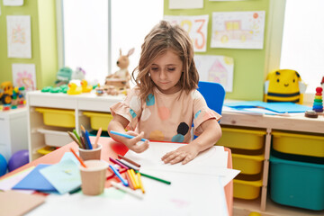 Adorable hispanic girl student drawing on paper sitting on table at kindergarten