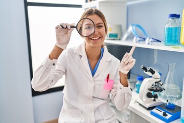Young blonde woman working at scientist laboratory using magnifying glass smiling happy pointing with hand and finger to the side