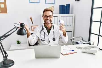 Middle age doctor man holding thermometer at the clinic celebrating victory with happy smile and winner expression with raised hands
