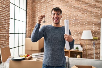 Young hispanic man holding keys of new home and blueprints sticking tongue out happy with funny...