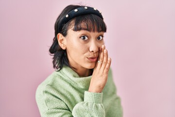 Young beautiful woman standing over pink background hand on mouth telling secret rumor, whispering malicious talk conversation