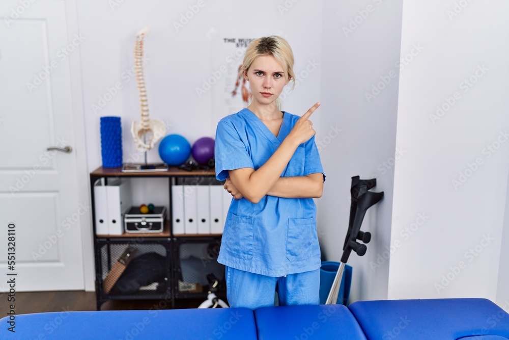 Sticker Young physiotherapist woman working at pain recovery clinic pointing with hand finger to the side showing advertisement, serious and calm face