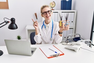 Young doctor woman holding model of human anatomical skin and hair at the clinic smiling happy pointing with hand and finger to the side