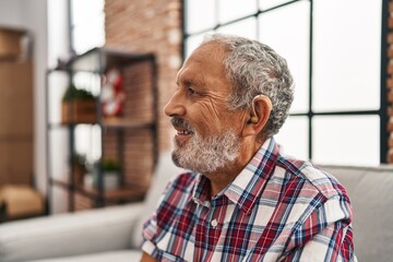 Senior grey-haired man using hearing aid sitting on sofa at home