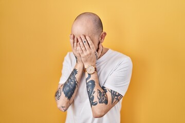 Young hispanic man with tattoos standing over yellow background with sad expression covering face...