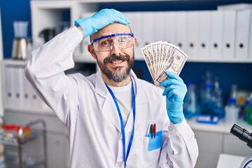 Young hispanic man working at scientist laboratory holding money stressed and frustrated with hand on head, surprised and angry face