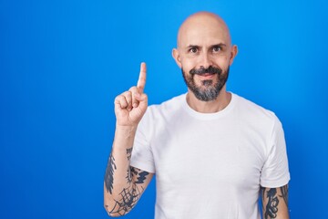 Hispanic man with tattoos standing over blue background showing and pointing up with finger number one while smiling confident and happy.