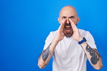 Hispanic man with tattoos standing over blue background shouting angry out loud with hands over mouth