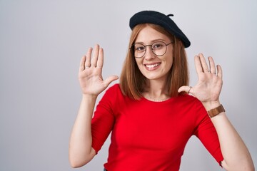 Young redhead woman standing wearing glasses and beret showing and pointing up with fingers number ten while smiling confident and happy.